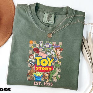 a green t-shirt with a graphic of characters from the Toy Story movie, including Woody, Buzz Lightyear, and other beloved characters. The t-shirt is laid out on a white surface, with a hat and some leaves visible in the background.