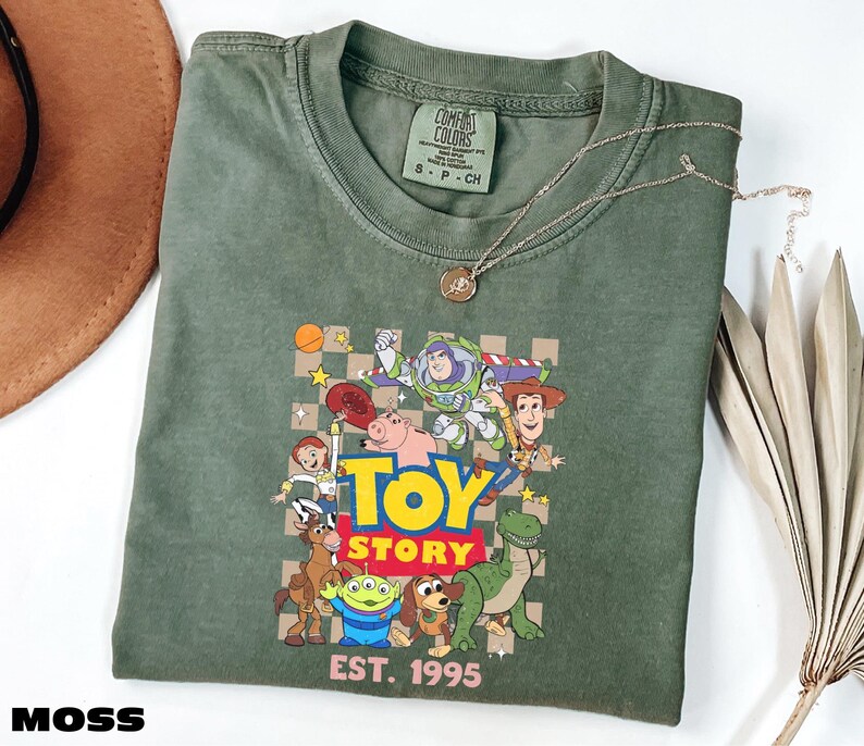 a green t-shirt with a graphic of characters from the Toy Story movie, including Woody, Buzz Lightyear, and other beloved characters. The t-shirt is laid out on a white surface, with a hat and some leaves visible in the background.