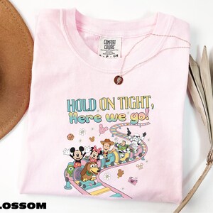 A pink t-shirt with a cartoon character design and text, along with a hat and a plant, is displayed on a white surface.