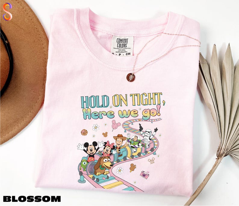 A pink t-shirt with a cartoon character design and text, along with a hat and a plant, is displayed on a white surface.