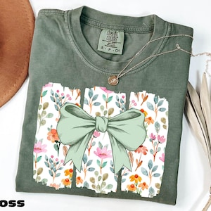 a green t-shirt with a bow design, placed on a white surface alongside a brown hat and some dried flowers.