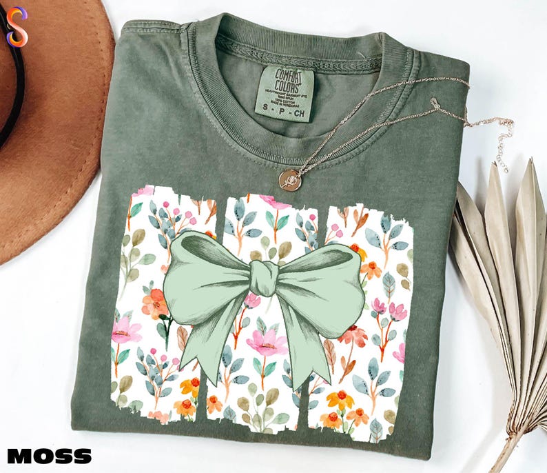 a green t-shirt with a bow design, placed on a white surface alongside a brown hat and some dried flowers.