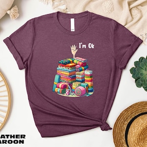 May include: Heather maroon t-shirt with a graphic of a pile of colorful fabric scraps and the text "I'm Ok".