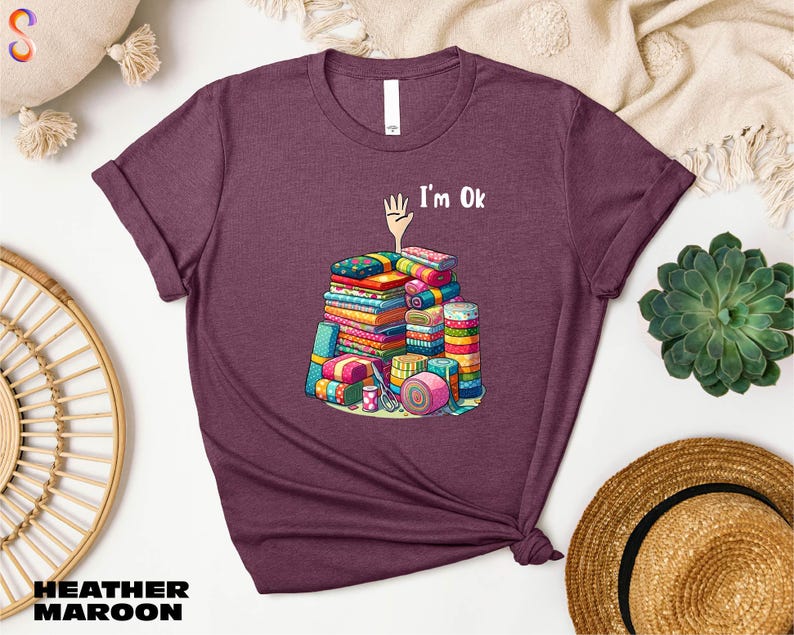 May include: Heather maroon t-shirt with a graphic of a pile of colorful fabric scraps and the text "I'm Ok".