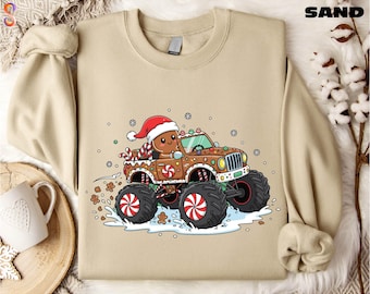 Könnte beinhalten: Sandfarbenes Sweatshirt mit einem Lebkuchenmann, der einen Lebkuchen-Monster-Truck fährt. Der Truck hat große schwarze Reifen mit roten und weißen Akzenten, und der Lebkuchenmann trägt einen Weihnachtsmannhut. Schneeflocken und Lebkuchenmänner sind verstreut. Das Wort 'SAND' ist oben.
