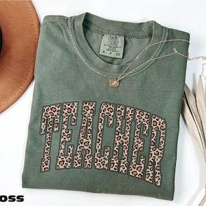 Puede incluir: Una camiseta verde musgo con la palabra "TEACHER" en un diseño de estampado de leopardo. La camiseta tiene cuello redondo y mangas cortas. Un collar dorado con un colgante está drapeado sobre la camiseta. La palabra "MOSS" está en la parte inferior izquierda.
