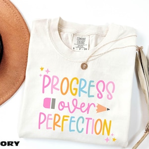 a white t-shirt with the text "PROGRESS OVER PERFECTION" printed on it, along with a pencil and a fan.