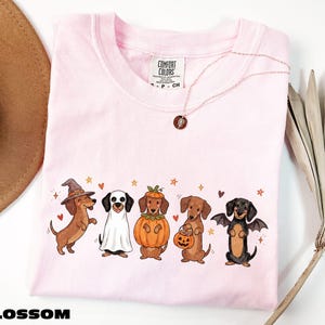A pink t-shirt with a graphic of five dogs dressed in costumes, including a witch, a pumpkin, a bat, a dog in a skeleton costume, and a dog in a skeleton costume holding a pumpkin. The t-shirt is displayed on a white background, with a brown hat and a plant visible in the background.