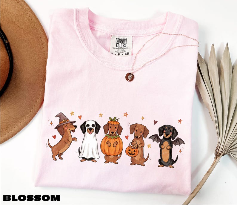 A pink t-shirt with a graphic of five dogs dressed in costumes, including a witch, a pumpkin, a bat, a dog in a skeleton costume, and a dog in a skeleton costume holding a pumpkin. The t-shirt is displayed on a white background, with a brown hat and a plant visible in the background.