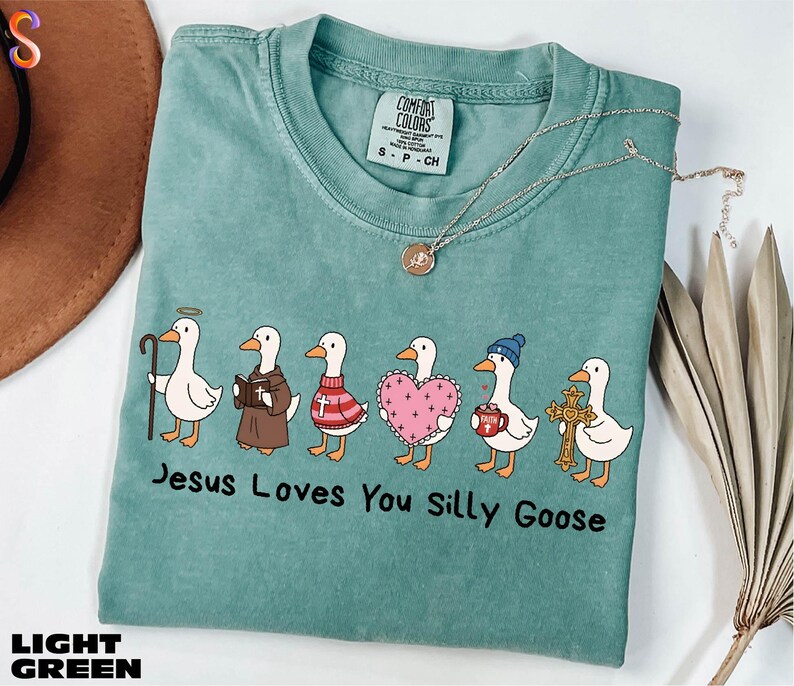 A light green t-shirt with a graphic design of six cartoon ducks, each with a unique outfit and accessory, arranged in a row. The text below the image reads "Jesus Loves You Silly Goose".