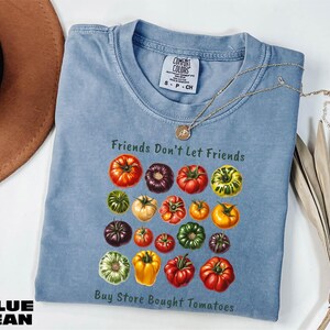 A blue denim t-shirt with a graphic of various colored tomatoes, along with the text "Friends Don't Let Friends" and "Buy Store Bought Tomatoes".