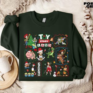 a green sweatshirt with a collage of various characters from the Toy Story franchise, including Woody, Buzz Lightyear, and other beloved characters, arranged in a festive manner. The sweatshirt is displayed on a bed with a white blanket and a snowflake decoration, creating a cozy and holiday-themed setting.
