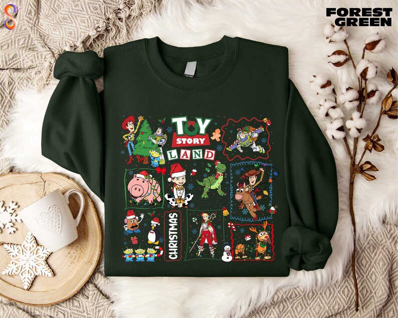 a green sweatshirt with a collage of various characters from the Toy Story franchise, including Woody, Buzz Lightyear, and other beloved characters, arranged in a festive manner. The sweatshirt is displayed on a bed with a white blanket and a snowflake decoration, creating a cozy and holiday-themed setting.