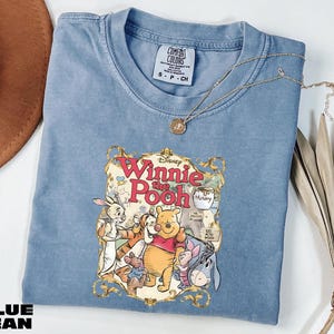A blue t-shirt with a Winnie the Pooh graphic and a necklace are displayed on a white surface.