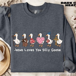 A gray sweatshirt with a graphic design of six cartoon ducks, each with a different outfit and accessory, arranged in a row. The text below the ducks reads "Jesus Loves You Silly Goose".