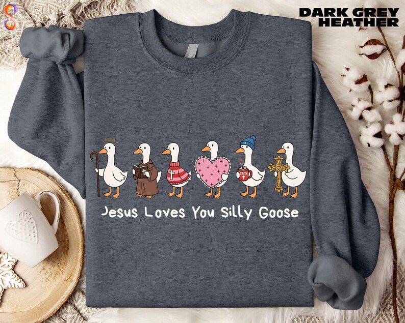 A gray sweatshirt with a graphic design of six cartoon ducks, each with a different outfit and accessory, arranged in a row. The text below the ducks reads "Jesus Loves You Silly Goose".