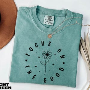 Peut inclure: T-shirt vert clair Comfort Colors avec le texte "FOCUS ON THE GOOD" en noir. Un dessin de marguerite est au centre du texte. Un collier doré est drapé sur le t-shirt.