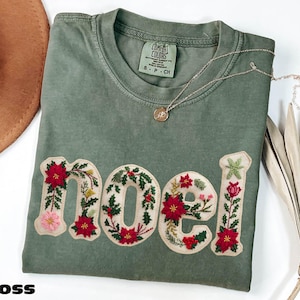 May include: Moss green t-shirt with the word "NOEL" embroidered with red poinsettias, green holly, and other floral accents. A gold necklace with a pendant rests on the shirt. The shirt is a Comfort Colors brand.