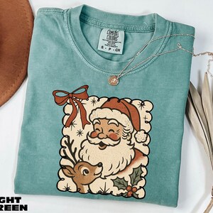 A turquoise t-shirt with a Santa Claus and reindeer graphic, along with a necklace and a hat, are displayed on a white surface.