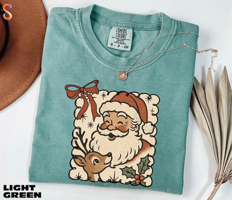 A turquoise t-shirt with a Santa Claus and reindeer graphic, along with a necklace and a hat, are displayed on a white surface.