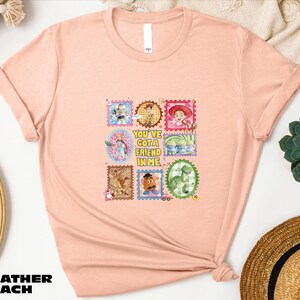 A pink t-shirt with a colorful graphic design featuring various cartoon characters and text, placed on a white surface alongside a straw hat and other decorative items.