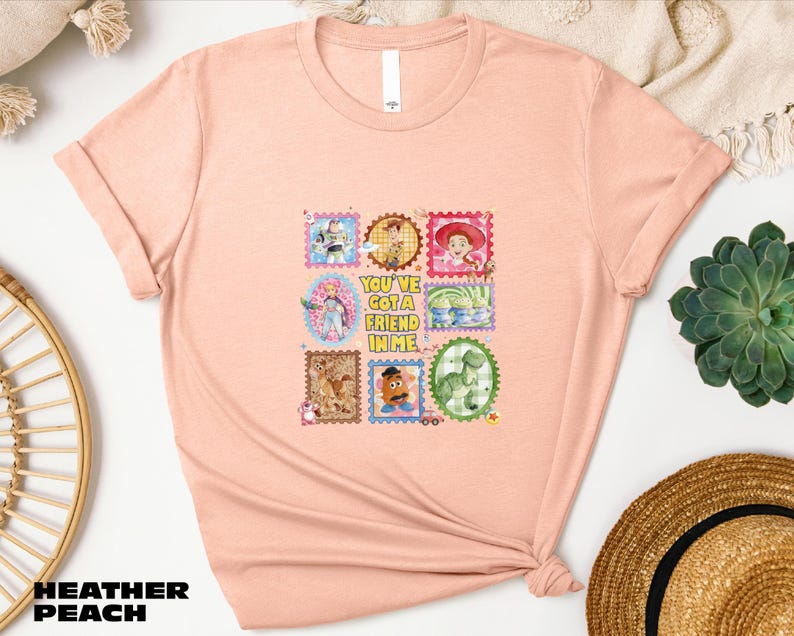 A pink t-shirt with a colorful graphic design featuring various cartoon characters and text, placed on a white surface alongside a straw hat and other decorative items.