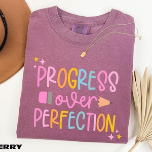 a purple t-shirt with the text "PROGRESS OVER PERFECTION" printed on it, along with a pencil and a fan.