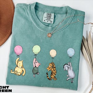A light green t-shirt with a design of Winnie the Pooh and his friends holding balloons, along with a necklace and a hat.
