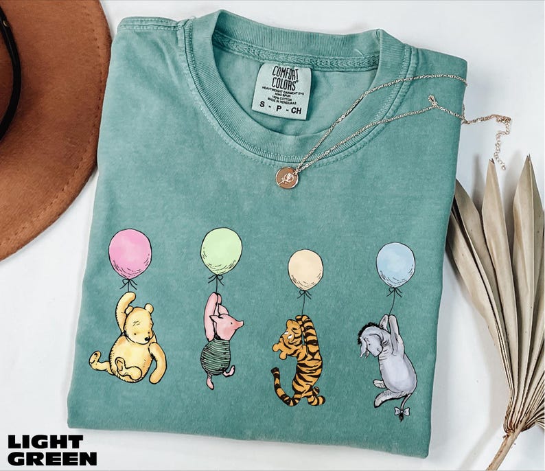 A light green t-shirt with a design of Winnie the Pooh and his friends holding balloons, along with a necklace and a hat.