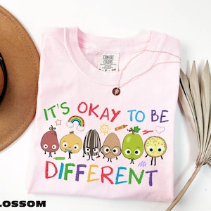A pink t-shirt with a colorful graphic design of cartoon characters and the text "It's OKAY TO BE DIFFERENT" printed on it.