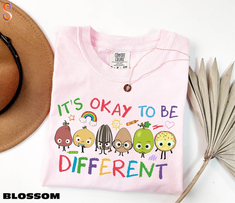A pink t-shirt with a colorful graphic design of cartoon characters and the text "It's OKAY TO BE DIFFERENT" printed on it.