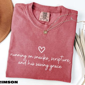May include: A crimson short-sleeved t-shirt with a crew neckline and a Comfort Colors label. The front features a white hand-drawn heart and the text: running on snacks, scripture, and his saving grace. A gold-toned necklace rests on the fabric.