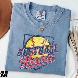 A blue softball-themed t-shirt with the text "SOFTBALL MAMA" printed on it, along with a necklace and a brown hat.