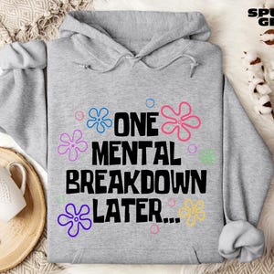 May include: A light gray hooded sweatshirt featuring the black text ONE MENTAL BREAKDOWN LATER... surrounded by colorful flower outlines in blue, pink, purple, green, and yellow, with the words SPORT GREY printed in the top right corner.