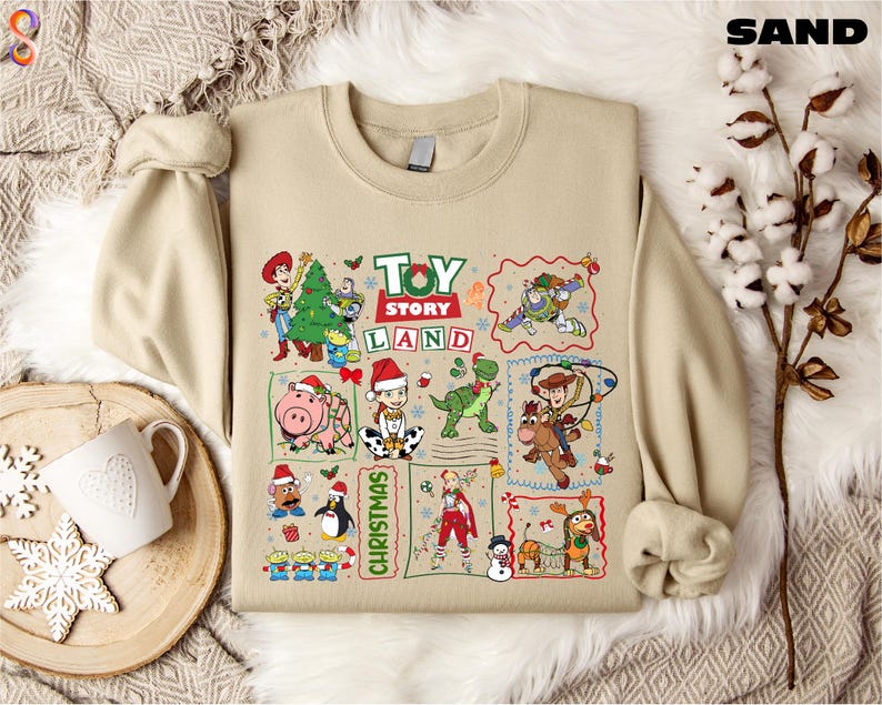 a beige T-shirt with a collage of various cartoon characters and scenes from the Toy Story Land movie franchise, including characters like Woody, Buzz Lightyear, and a snowman. The T-shirt is displayed on a bed with a white blanket and a mug, creating a cozy and festive atmosphere.