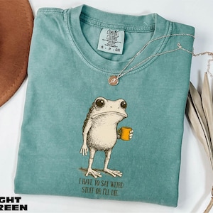 May include: Light green Comfort Colors t-shirt featuring a whimsical illustration of a frog holding a yellow mug. The frog has large eyes and is standing upright. The text "I HAVE TO SAY WEIRD STUFF OR I'LL DIE" is printed below the frog. A silver necklace is draped across the shirt.