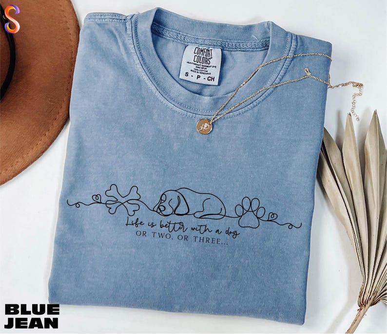 A blue denim t-shirt with a graphic design and text printed on it, along with a necklace and some dried leaves.