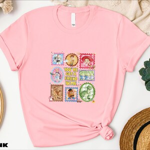 A pink t-shirt with a colorful graphic design featuring various cartoon characters and text, placed on a white surface alongside other items such as a hat and a plant.