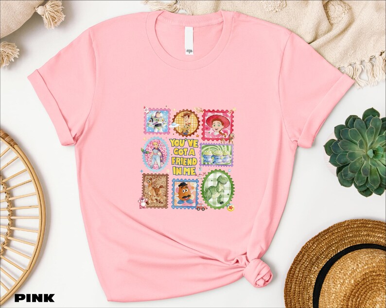A pink t-shirt with a colorful graphic design featuring various cartoon characters and text, placed on a white surface alongside other items such as a hat and a plant.