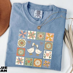 A blue denim t-shirt with a colorful quilt-like design featuring two white ducks, flowers, and geometric shapes. The t-shirt is placed on a white surface, with a brown hat and some dried leaves nearby.