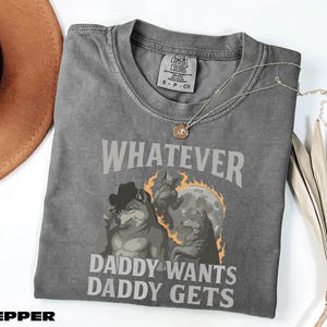 May include: A grey Comfort Colors t-shirt featuring the text "WHATEVER DADDY WANTS DADDY GETS" and a wolf graphic. The shirt has a crew neck and short sleeves. A gold necklace is draped across the shirt.