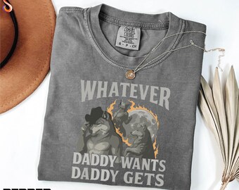 May include: A gray Comfort Colors t-shirt with the text "WHATEVER DADDY WANTS DADDY GETS" and a graphic of wolves. The shirt has a crew neck and short sleeves. A gold necklace is draped across the shirt.