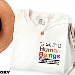 May include: Ivory t-shirt with a rainbow colored graphic that says "Human Beings" with the text "100% Organic" and "Colors may vary."