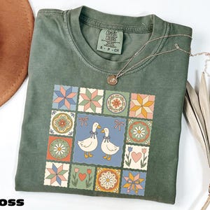 a green t-shirt with a colorful, patterned design featuring two cartoon ducks, flowers, and stars. The t-shirt is placed on a white surface, and there is a brown hat and some dried leaves nearby.