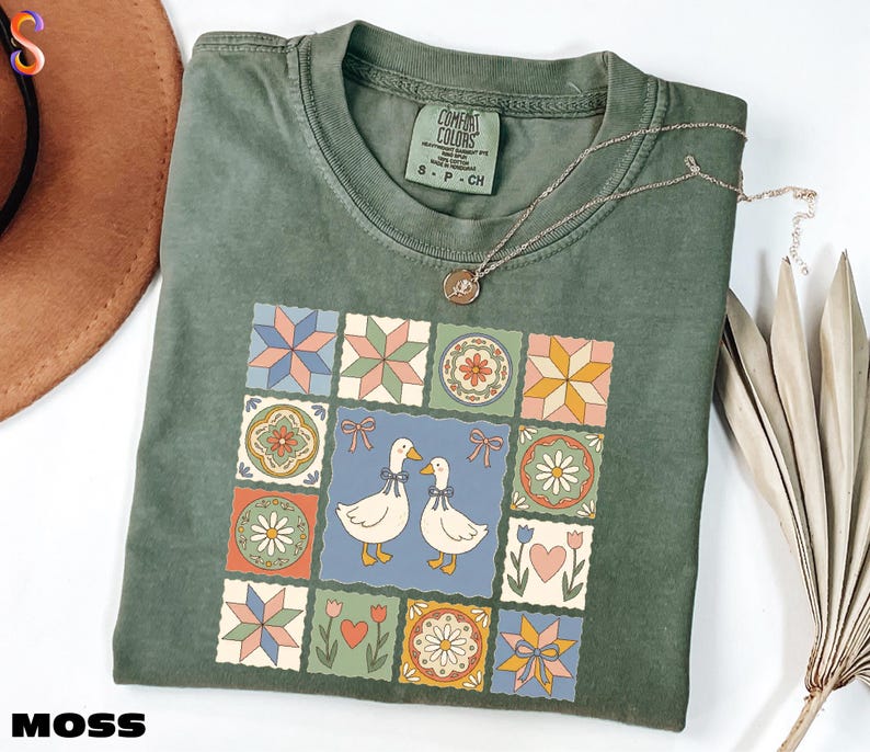 a green t-shirt with a colorful, patterned design featuring two cartoon ducks, flowers, and stars. The t-shirt is placed on a white surface, and there is a brown hat and some dried leaves nearby.