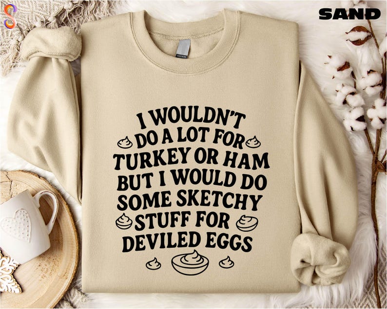 A beige sweatshirt with a humorous text printed on it, along with a mug and some decorative elements like a branch and a bowl.
