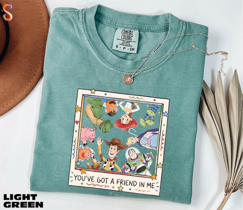 A green t-shirt with a colorful graphic of characters from the Toy Story movie, along with the text "You've Got a Friend in Me" printed on it. The t-shirt is placed on a white surface, and there is a brown hat and some dried leaves nearby.