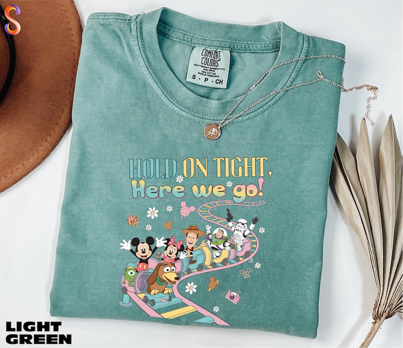 A light green t-shirt with a colorful graphic of Disney characters on a roller coaster, along with the text "HOLD ON TIGHT, HERE WE'VE GO!" The t-shirt is displayed on a white surface, with a brown hat and some dried leaves in the background.