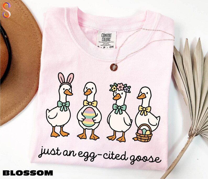 A pink t-shirt with a cartoon illustration of four cartoon ducks wearing Easter-themed accessories, accompanied by the text "just an egg-cited goose".