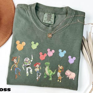 a green t-shirt with a design of various cartoon characters, including a character holding a balloon, a character holding a balloon, and several other characters, all depicted in a playful and colorful manner.